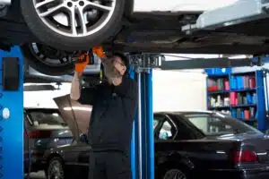 A mechanic wearing orange gloves and a black hoodie uses a power tool to repair a car lifted on a hydraulic hoist.
