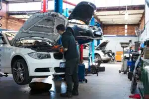 A mechanic in a black hoodie and orange gloves works on a lifted BMW, with multiple cars being repaired in the background.
