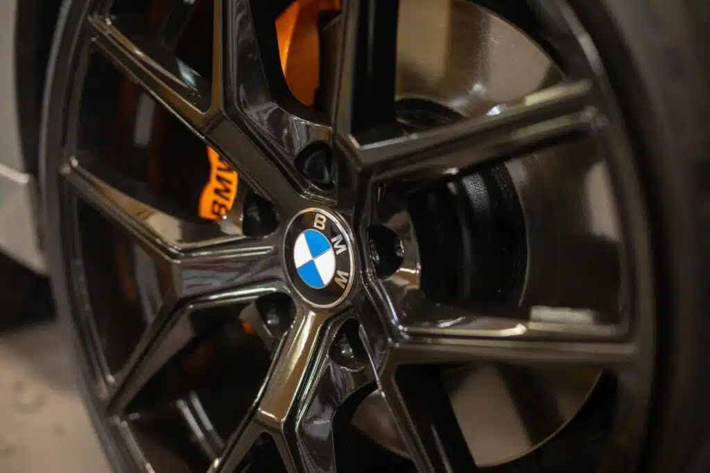 A close-up of a BMW alloy wheel with a glossy black finish and an orange brake caliper visible behind it.