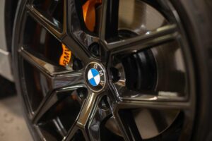 A close-up of a BMW alloy wheel with a glossy black finish and an orange brake caliper visible behind it.