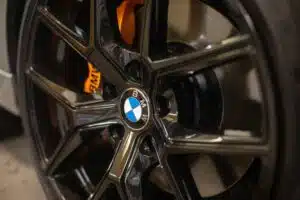 A close-up of a BMW alloy wheel with a glossy black finish and an orange brake caliper visible behind it.