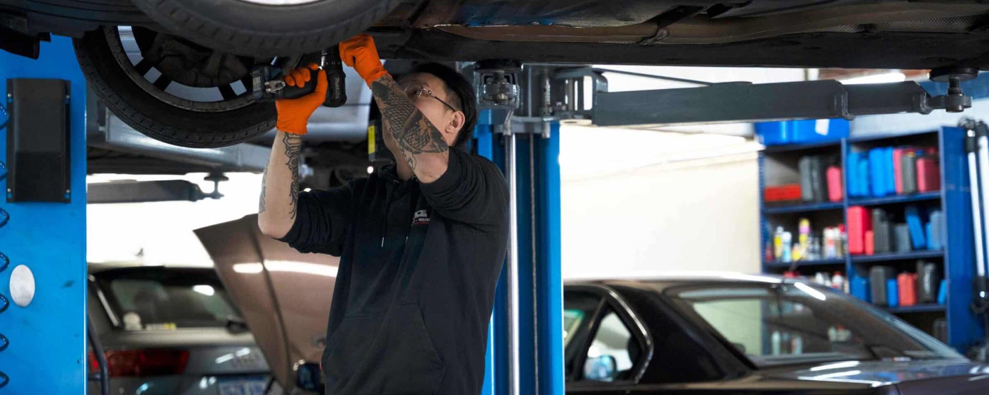 A mechanic wearing orange gloves and a black hoodie uses a power tool to repair a car lifted on a hydraulic hoist.